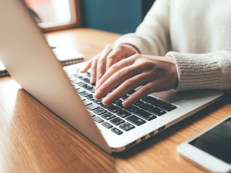 A close up of hands typing at a laptop on a wooden desk. 